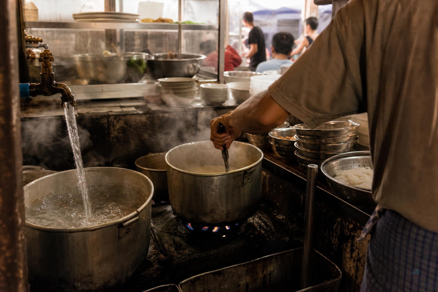 Busy Myanmar restaurant kitchen during service — pots filling, staff working at speed under warm light